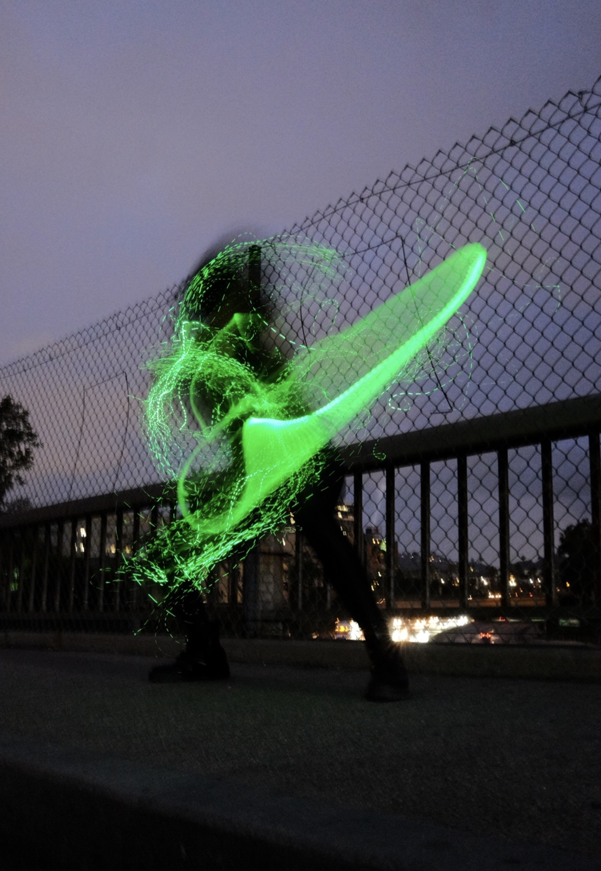 Long-exposure photo of Luna spinning a green LED whip on a city bridge at dusk